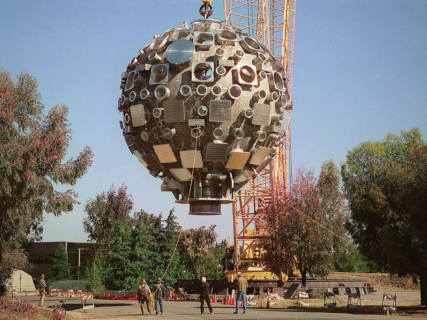 The spherical target chamber of the National Ignition Facility hangs from a construction crane above workers